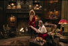A warmly lit room with a fireplace, bookshelves, and vintage decor resembles a scene from The Christmas Chronicles (2018) DVD: a woman in a red coat stands by the fire, an older man reads nearby, with a dog and elves resting close.
