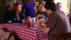 Three people enjoy food and drinks at a cozy, sunlit restaurant table with a red and white checkered cloth, chatting about vintage films while "Dear Prudence (2009) DVD" sits among them, adding to their cheerful gathering.