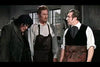 Three men in period clothing and aprons stand in a workshop, evoking the eerie mood of a classic horror film—reminiscent of scenes from the Blood of the Vampire DVD (1958)—amid tools and windows, with one looking down sadly.