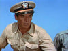 A man in a tan military uniform and white naval officer’s cap stands outdoors against a clear blue sky, evoking the determined spirit seen in classic films like The Sharkfighters (1956) DVD.