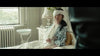 A woman with a bandaged head and arm sits in bed, glancing at someone nearby. On the windowsill beside her rests a Godzilla Minus One (2023) DVD, its cover visible in the softly lit, patterned room.