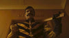 A man in a yellow and black striped uniform with a large mustache and gloves holds a bat with a stern look in a dimly lit room, as seen in the supernatural film "Haunting of Queen Mary (2023)" on DVD.