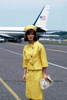 A woman in a yellow suit and hat stands on an airport runway holding a white clutch, evoking the elegance of Jacqueline Kennedy, with an airplane featuring a U.S. flag in the background—an image reminiscent of scenes from the "Jacqueline Bouvier Kennedy DVD (1981).