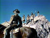 A person in a cowboy hat sits on a horse, gazing at spear-carrying figures in traditional dress—evoking scenes from rare westerns—on rocky terrain under blue sky, much like moments found in the "Kentucky Rifle (1955) DVD.