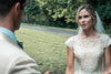 A bride in a white lace dress and veil stands outdoors, gazing at a man in a light suit—just like a scene from the Murder at the Mansion (2018) DVD. Lush trees and grass complete the backdrop.
