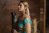 A blonde woman stands indoors by a wooden wall wearing a colorful plaid dress and necklace. "Karl + Nicole" is carved into a beam—evoking scenes from the Murder at the Mansion (2018) DVD. Straw can be seen in the background.