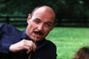 A man with a bald head and mustache, dressed in a dark shirt, reclines outdoors with a piece of straw in his mouth. Behind him, a blurred green lawn and dark wooden fence enhance the relaxed vibe—like someone unwinding after adding “Love, Murder and Deceit (1997) DVD” to their rare movie collection.