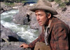 A man in a plaid jacket and cowboy hat kneels by a rushing river on rugged terrain with greenery, evoking the adventurous spirit of "The Naked Spur (1953) DVD.