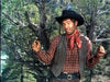 A man dressed in James Stewart’s western style stands outdoors, wearing a black hat, red scarf, plaid shirt, and vest—similar to his look in "The Naked Spur (1953)," now available on DVD.