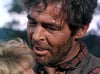 A rugged man with messy dark hair and a scruffy beard in a plaid shirt speaks to a blonde woman outdoors, echoing the classic western atmosphere of "The Naked Spur (1953) DVD." Blurred greenery adds to the timeless scene.