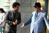A man in a dark suit talks to an older man in a light blue suit and black hat outside an ornate building, discussing the Nothing in Common (1986) DVD as people walk by in the background.