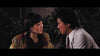 A woman in colorful clothing and a man in a white shirt and tie sit closely together outdoors at night, gazing at each other with intense expressions, as if they were characters straight out of “Oily Maniac (1976),” a cult classic now available on DVD.