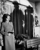 A woman holding a gun stands by a table in a vintage room, looking to the side. Ominous shadows loom on the curtains behind her, evoking the tense, suspenseful atmosphere of a scene straight out of Out of the Past (1947), now available on DVD.