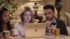 Three people sit closely together at a table, looking intently at a laptop screen. Holiday mugs and a decorated Christmas tree are visible in the background, creating a warm, festive atmosphere—perhaps they’re enjoying their holiday tradition by watching “A Christmas Prince: The Royal Wedding (2018) DVD” or reminiscing over other classic movies on DVD.