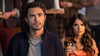 A man and a woman look ahead with serious expressions at a social gathering. The man wears a dark jacket and beaded necklace, while the woman in orange holds a glass. Blurred partygoers fill the background, evoking the atmosphere of “Love Hard (2021) DVD,” one of those rare holiday rom-coms that captures fleeting moments at festive gatherings.