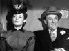 A woman in a patterned dress and feathered hat sits beside a smiling man in a suit and fedora, both dressed in vintage 1940s attire—evoking the timeless elegance featured in *The Ghost and Mrs. Muir (1947)* DVD.