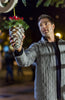 A man in a gray quilted jacket holds up a sparkly pine cone ornament with red ribbon, standing outdoors at night under festive decorations—just like a scene from “Magic Stocking (2015)” on DVD.