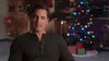 A man in a dark henley shirt sits indoors with a decorated Christmas tree and wrapped gifts blurred in the background, capturing the cozy holiday atmosphere reminiscent of “Magic Stocking (2015)” on DVD.