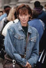 A woman with short brown hair wearing a blue denim jacket stands among a crowd of people outdoors, gazing toward the camera with a neutral expression, as if she’s just discovered a hidden stall selling rare hard-to-find movies like The Counterfeit Contessa (1994) on DVD.