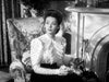 A woman evocative of Gene Tierney in Victorian-era attire sits thoughtfully beside a fireplace on a floral-patterned armchair, reminiscent of a scene from “The Ghost and Mrs. Muir (1947)” DVD, captured in striking black and white.