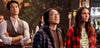 Three people stand indoors, looking intently ahead, as if caught in a scene from the “Love Hard (2021) DVD.” The man on the left wears a vest, the person in the center sports glasses and a puffy jacket, and the woman on the right wears a red plaid shirt over a camp T-shirt.