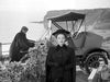 A woman in dark Victorian-era clothing stands by a cliffside road, capturing the timeless elegance found in The Ghost and Mrs. Muir (1947) DVD, while a man in similar attire leans on a fence near an old-fashioned car. The ocean and rocky cliffs stretch behind them, evoking haunting scenes akin to those once graced by stars like Gene Tierney.