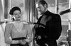 A woman in a high-neck blouse stands beside a man dressed in dark clothing, the pair positioned indoors near a curtained window and engaged in an intense conversation. Captured in black and white, the scene evokes the timeless atmosphere of “The Ghost and Mrs. Muir (1947) DVD.”.