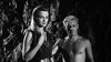 A woman in a dark dress and necklace stands next to a shirtless, curly blond man in a rocky cave—just like a scene from the rare cult classic "Saga of the Viking Women (1957) DVD," both staring intently at the camera.