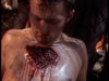 A person sits shirtless against a fence, looking down at a large, bloody wound on their chest, with blood dripping down their torso—a scene reminiscent of the intense battles seen in "Shootfighter" (1993), now available on DVD.