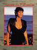 A woman stands on the beach in a black swimsuit and sheer cover-up, wet hair framing her serious expression. Above her: "Trouble in Paradise (1989) DVD"—the classic film starring Raquel Welch and Jack Thompson, now available.