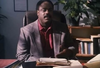 A man in a gray blazer sits at a desk with paperwork and books, perhaps cataloging the “With Murder in Mind (1992) DVD,” with shelves and potted plants in the background.