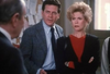 A man in a suit and a woman with curly blonde hair and a red top stand together indoors, appearing concerned as they listen to someone offscreen. Echoing the suspenseful atmosphere of classic films like With Murder in Mind (1992) on DVD, papers are pinned to a board in the background.