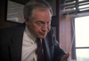 An older man in a suit leans forward in conversation near a window with blinds, his serious expression evoking the intense moments seen in films like “With Murder in Mind (1992)” on DVD.