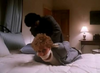 A woman with curly blonde hair is lying on a bed, her hands and feet bound behind her back and a gag around her mouth. A person stands behind her in a dimly lit bedroom, where posters of rare DVDs—including “With Murder in Mind (1992)”—and images of Elizabeth Montgomery decorate the walls, evoking an air of vintage intrigue.