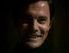 A man with dark hair and pale skin smiles, revealing sharp vampire fangs, his face partially lit against a dark background—reminiscent of scenes from the Count Dracula (1977) DVD.