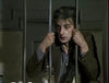 A man with messy hair and a surprised look grips jail bars, wearing a brown jacket and gray shirt. On the table in front of him is the "Count Dracula (1977) DVD" among assorted items.