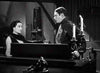 A woman and a man stand facing each other in a dimly lit room, capturing the mysterious atmosphere featured in classic horror films. The man stands behind a piano topped with two flickering candles, while the woman regards him with a serious expression—reminiscent of scenes from Dracula’s Daughter (1939), now available on DVD.