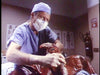 A surgeon wearing scrubs and a mask operates on a patient’s severely injured, bloody arm in a hospital room—a scene that evokes the intense atmosphere of cult horror classics like those featured in *Maniac Cop DVD (1988)*. Medical posters and a fire extinguisher are seen in the background, reminiscent of moments found in rare movies on DVD such as this.