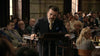 A man in a dark suit leans forward at a courtroom podium, gazing intensely ahead—a gripping scene that recalls the suspense of Find Me Guilty (2006) DVD—as people watch from the background.