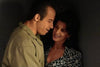 A man in a khaki shirt stands close to a woman with dark hair in a patterned blouse, sharing an affectionate moment in dim light—like a scene from the Find Me Guilty (2006) DVD.