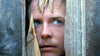 A person with blue eyes peers through a jagged hole in a wooden door, looking intently out from between the broken wood panels—reminiscent of a suspenseful scene from “The Frighteners (1996)” DVD, where curiosity and tension hold you captive throughout the film.