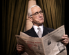 An older man in a suit and glasses sits in front of draped curtains, holding and reading a newspaper with a slight smirk on his face—much like a collector examining reviews for rare classic movies on DVD or enthusiasts discussing the acclaimed Give’em Hell Harry! DVD (1975), the celebrated biographical play about Harry S Truman.