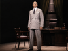 An older man in a gray suit stands confidently in a dimly lit office, with a wooden desk, leather chair, and dark green curtains in the background—a scene evocative of “Give ’em Hell Harry! DVD (1975).”.