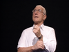 An older man wearing glasses and a white shirt stands against a dark background, gazing upward with a thoughtful expression and one hand clenched before him—striking a pose reminiscent of pivotal moments captured in the “Give’em Hell Harry! DVD (1975),” the biographical play about Harry S Truman.