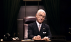 An older man with white hair and glasses sits at a desk in a dimly lit office, dressed in a suit and tie. He holds a pen, while a nameplate, horseshoe, and vintage phone rest on the desk—evoking moments from Give’em Hell Harry! DVD (1975), the celebrated Harry S Truman biographical play available alongside other rare classic movies on DVD.