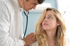 A doctor in a white coat examines a young woman with a stethoscope, reminiscent of scenes from "The Good Doctor (2011)" DVD, as she sits and looks up at him during the medical checkup.