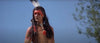 A person with long dark hair, red and white face paint, and a feather headdress stands outdoors against a blue sky—evoking scenes from the "Grayeagle (1977) DVD" and classic Native American frontier adventures.