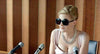 Wearing large black sunglasses, a silver necklace, and a light pink dress, a woman sits confidently with two microphones before a wooden panel backdrop—capturing the captivating aura found in "Nelly (2016)" on DVD.