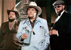 Three men stand together indoors: one, reminiscent of Willie Nelson in a light blue cowboy suit and hat while holding a drink, is flanked by two men in brown suits and hats—as if ready to star in *Songwriter (1984) DVD*.
