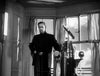 A man in dark clothing stands in a bright room with large windows and a nautical instrument, possibly a ship’s bridge, sunlight streaming in and clouds visible outside—evoking the haunting atmosphere of “The Ghost and Mrs. Muir (1947)” on DVD, where stars like Gene Tierney once graced the screen.